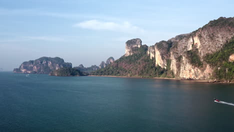 Long-tail-boat-cruising-on-sea-below-mountain-cliff-coast-of-Thailand