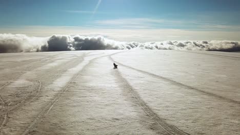 Aerial-panoramic-view-of-a-person-riding-a-snowmobile-on-a-glacier-in-Iceland,-over-the-clouds,-on-a-sunny-day