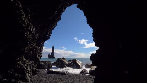 Waves-crash-on-large-black-rocks-with-stoic-rock-spire-in-ocean,-black-beach-Iceland