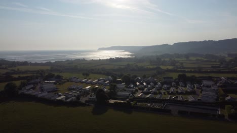 Aerial-rising-view-Welsh-caravan-park-at-sunrise-overlooking-shimmering-coastal-bay-and-rolling-countryside-hills