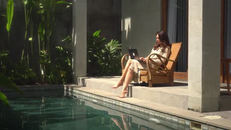 Expat-woman-working-online-in-her-luxury-home-with-swimming-pool-during-sunny-day-and-using-laptop-computer-to-work-from-home-early-morning-in-Bali,-Indonesia