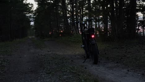 Electric-bicycle-parked-on-country-road-during-sunset-with-red-backlight-on,-trees-moving-in-wind