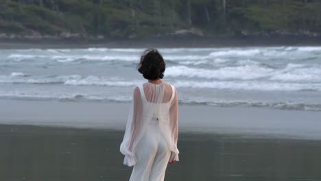 Mujer-Vestida-De-Blanco-Caminando-Descalza-En-La-Playa-Con-Olas-ásperas-En-El-Fondo