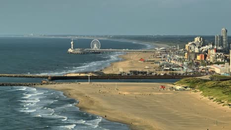 Aerial-wide-shot-of-beautiful-sandy-beach-and-ferris-wheel-on-jetty-with-Den-Haag-City-on-the-right-side