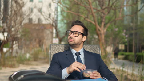 Pensive-millennial-businessman-eating-nuts-in-park-outdoors-enjoying-lunch-break