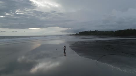 Aerial-tracks-Asian-man-riding-motorcycle-on-wide-grey-ocean-beach