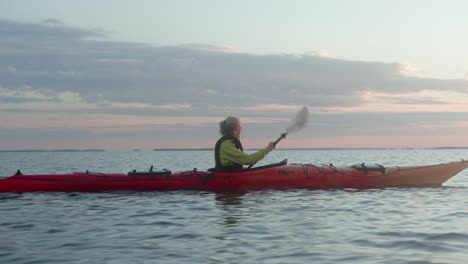 Young-Blonde-Woman-Paddling-in-a-Red-Sea-Kayak-on-the-open-Sea-in-Finland,-Vaasa,-Archipelago,-Beautiful-Summer-Sunset-Atmosphere,-Dawn,-Close-up