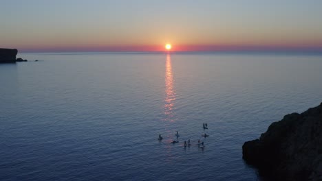 Drone-flying-over-paddle-boarders-with-the-reflection-of-sunset-beaming-across-the-water-in-Menorca,-Spain