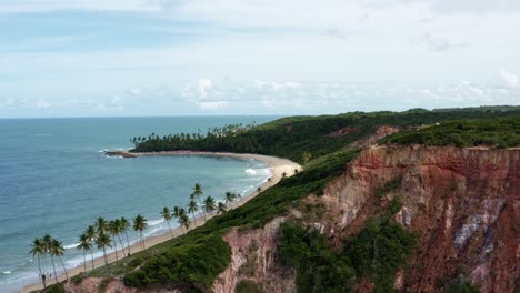 Left-trucking-aerial-drone-extreme-wide-shot-of-the-popular-tropical-Coquerinhos-beach-with-colorful-cliffs,-palm-trees,-golden-sand,-small-waves,-and-turquoise-water-in-Conde,-Paraiba,-Brazil