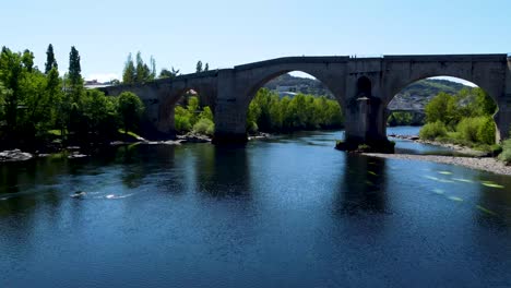 Low-angle-aerial-towards-and-through-ancient-Roman-bridge,-Ourense