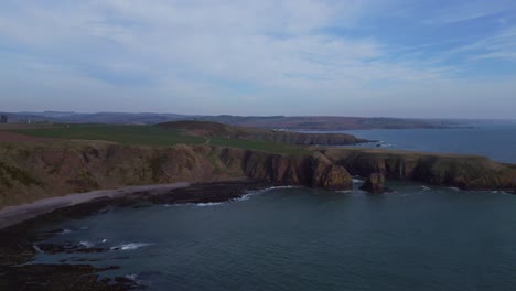 Aerial-drone-footage-gliding-over-Dunnottar-Castle,-soaring-above-the-ocean-towards-the-picturesque-beach-and-lush-green-clad-cliffs