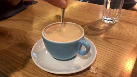 Close-up-view-of-stirring-creamy-cappuccino-with-a-tea-spoon-served-on-a-wooden-table