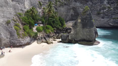 Aerial,-Diamond-shaped-Rock,-Palm-trees-and-People-swimming-at-Diamond-Beach-in-Nusa-Penida-Island