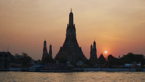 Vista-Del-Templo-Wat-Arun-En-Bangkok-Durante-Una-Impresionante-Puesta-De-Sol,-Con-Un-Ferry-Tradicional-Cruzando-El-Río-Chao-Phraya-Frente-A-él