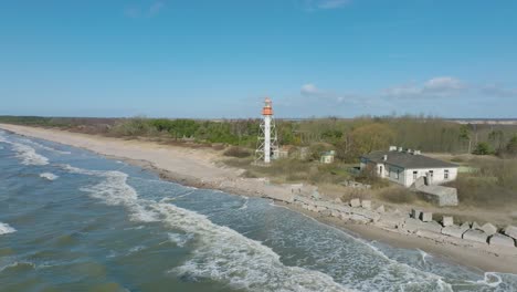 Aerial-establishing-view-of-white-colored-Pape-lighthouse,-Baltic-sea-coastline,-Latvia,-white-sand-beach,-large-waves-crashing,-sunny-day-with-clouds,-wide-ascending-drone-shot-moving-forward