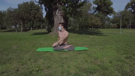 Beautiful-fit-young-man-with-mask-practice-yoga-in-the-park,-brunette-man-doing-exercise-and-spending-active-time