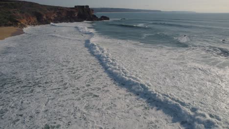 empty-beach-in-Nazare-near-a-massive-cliff-with-waves-gently-crashing-on-the-sand