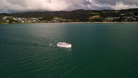 Beautiful-aerial-tracking-orbit-shot-of-boat-sailing,-reveal-of-small-beachfront-settlement-Langs-Beach,-New-Zealand