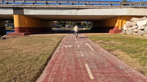 Old-man-pedals-on-bike-along-bike-path-on-sunny-day,-enters-below-bridge