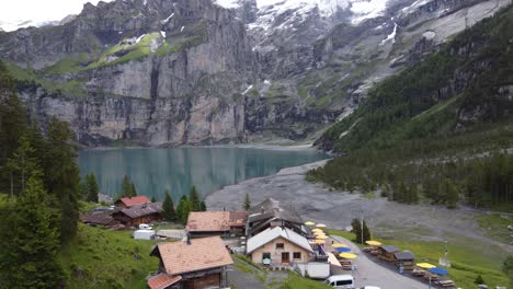 Toma-Aérea-Ascendente:-Vista-De-La-Cafetería-De-La-Montaña-Y-Las-Montañas-Gigantes-Bluemlisalp-Que-Rodean-El-Lago-Glaciar-Turquesa-Szure-Oeschinensee-En-Kandersteg,-Suiza