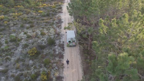 man-running-alongside-a-green-travel-camper-van-down-a-dirt-road-with-a-forest-next-to-them-at-sunset-in-nazare-portugal
