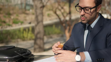 Young-bearded-businessman-eating-snacks-and-writing-notes,-working-with-papers-at-park-during-lunch-break
