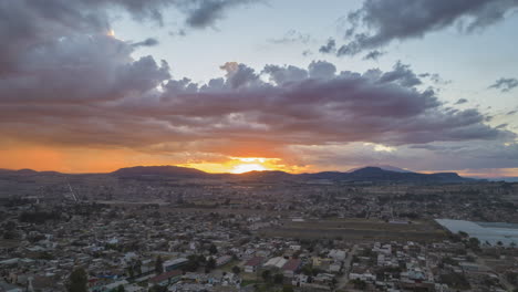 Captivating-hyperlapse-of-a-sunset-with-orange-hues-reflecting-on-clouds,-creating-a-beautiful-color-spectacle-as-the-sun-disappears-behind-mountains,-while-showcasing-the-village
