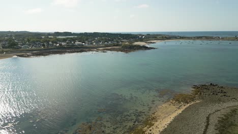 sweeping-drone-footage-of-Guernsey-coastline,-stunning-golden-beach-and-clear-calm-sea-with-boats-at-anchor-on-sunny-summer-day