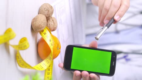 Vertical-video-of-Doctor-pointing-at-Smartphone-green-screen-with-Walnuts-and-Orange-fruit-on-table,-Healthy-diet