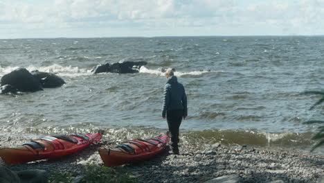 Wide-Shot-of-Blonde-Girl-Standing-on-the-beach-by-the-Sea,-Looking-out-to-the-waves,-Two-Kayaks-Laying-on-the-Beach,-Windy-Cold-Summer-Day-in-Finland,-Near-Vaasa