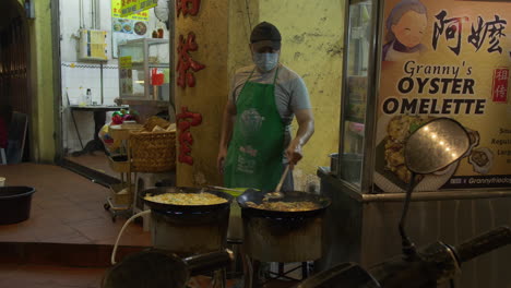 Un-Hombre-Vestido-Con-Delantal-Verde-Prepara-Comida-Callejera-Malaya-En-Wok,-Cocina-De-Gas-Caliente,-Mercado-Nocturno,-Comida-Callejera,-Malasia