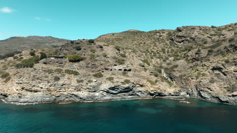 Aerial-view-towards-rustic-stone-mountainside-house-built-in-the-Cap-de-Creus-national-park-cliffs-overlooking-shimmering-emerald-water