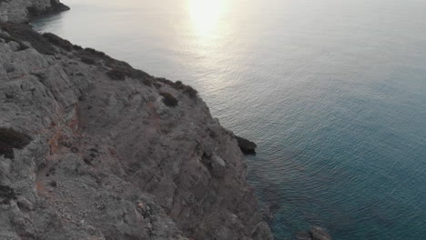 drone-captures-breathtaking-beauty-of-a-rocky-cliff-edge,-as-camera-soars-over-rugged-landscape-before-tilting-up-towards-horizon,-where-morning-sun-shines-over-endless-expanse-of-water