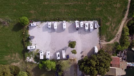Aerial-view-of-a-rectangular-parking-area-with-parked-campervans,-grass-all-around,-house-roofs-and-small-boats-in-the-lower-right