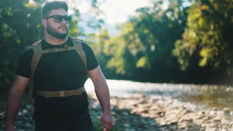 Young-male-hiker-with-sunglasses-walks-along-rocky-river-hot-summer-day,-towards-the-camera-direction-close-up-outdoor-activity-and-travel-in-nature