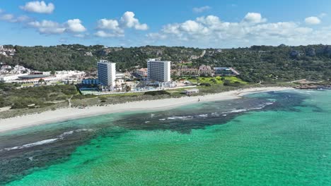 A-cinematic-aerial-view-of-Son-Bou-Beach-with-buildings-in-the-background-in-Menorca,-Spain
