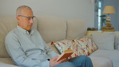 Hombre-Adulto-Leyendo-Un-Libro-En-Un-Sofá-Moderno-En-Casa,-Con-Una-Camisa-Azul-En-Un-Día-Soleado