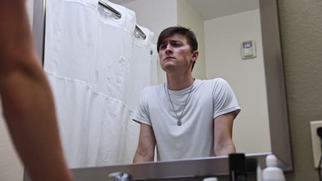Ein-Junger-Weißer-Mann-In-Einem-Weißen-T-Shirt-Geht-Deprimiert-Auf-Den-Badezimmerspiegel-Eines-Hotelzimmers-Zu-Und-Seufzt-Vor-Trauer-Im-Spiegelbild
