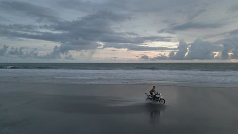 Man-rides-motorccle-on-sandy-ocean-beach-in-cloudy-morning-light