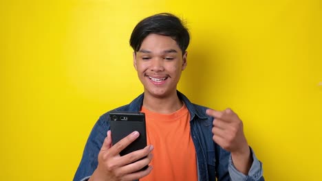 Excited-young-man-holding-mobile-phone,-showing-thumb-up-and-smiling,-pointing-finger-to-smartphone-isolated-on-yellow-background