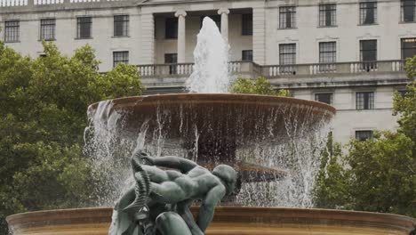 Cement-two-tiered-water-fountain-flowing-with-green-bronze-statue-in-foreground-and-white-urban-business-building-in-background,-London,-England,-static-slow-motion