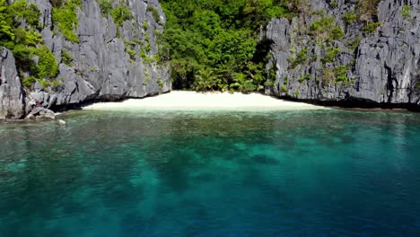 Aerial,-untouched-Hidden-Gem-of-a-white-sand-Beach-enclosed-by-lush-Karst-cliffs,-tropical-Jungle-and-turquoise-clear-water-on-Matinloc-Island,-El-Nido---Palawan