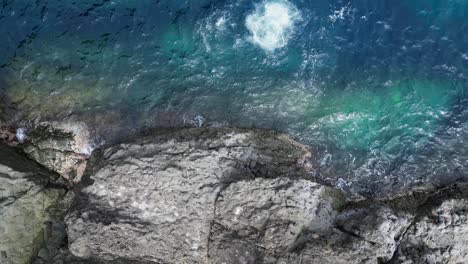 Aerial-Top-down-view-of-Male-Cliff-Diver-diving-into-turquoise-ocean-water-from-rocky-cliffside