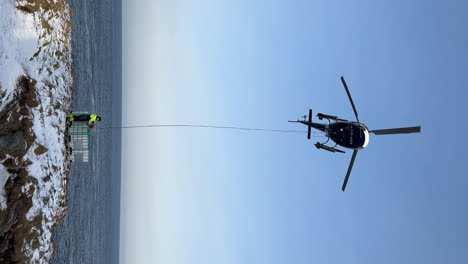 Vertical-shot-of-a-helicopter-with-a-worker-hooking-container-on-a-longline,-during-strong-wind-in-the-Arctic-Polar-circle