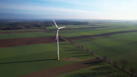 Flying-toward-spinning-wind-turbine-towering-by-countryside-road-against-plain-agricultural-farmland-in-Poland---aerial-flyover