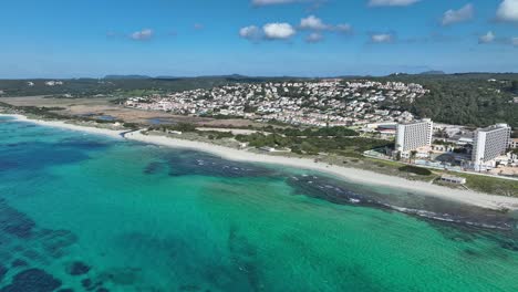 A-cinematic-aerial-view-of-Son-Bou-Beach-with-buildings-in-the-background-in-Menorca,-Spain