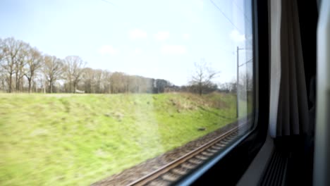 POV-Looking-Out-Train-Window-Going-Past-Bare-Trees-With-Clouds-And-Blue-Skies-Above-In-Background