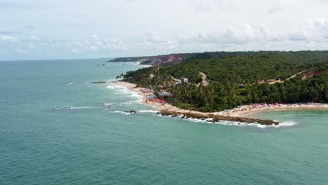 Rotating-aerial-drone-wide-shot-of-the-popular-tropical-Coquerinhos-beach-covered-in-umbrellas-with-tourists-swimming-in-a-natural-pool-from-a-reef-in-Conde,-Paraiba,-Brazil-on-a-warm-summer-day