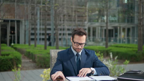 Caucasian-businessman-man-in-suit-doing-paperwork-in-a-park,-watching-phone-while-sitting-by-the-table-in-city-park-by-the-office---orbit-shot