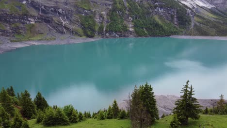 Aerial-drone-view-green-alpine-meadows-surrounded-by-Bluemlisalp-mountains,-pine-trees-overlooking-turquoise-azure-glacier-lake-Oeschinensee-in-Kandersteg,-Switzerland